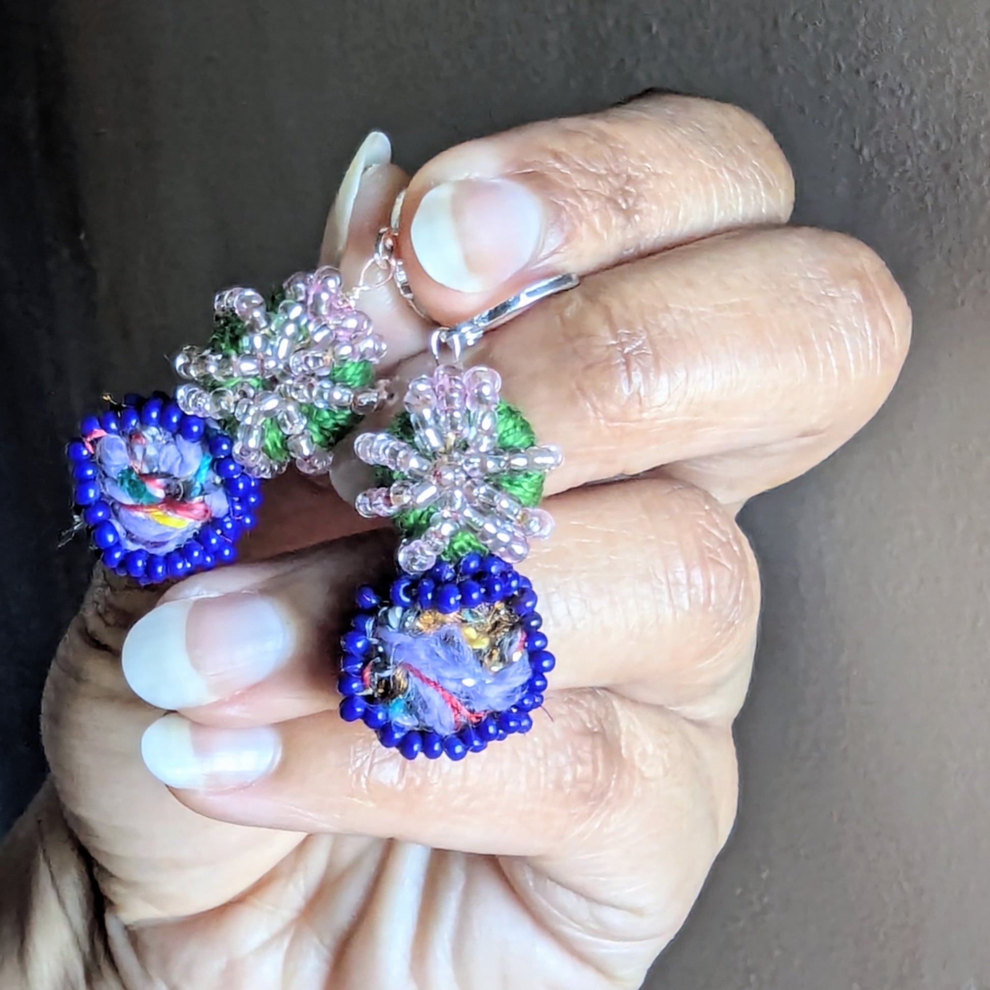 Hand holding Gaia earrings showing green crochet thread, blue bead frame, and pink glass seed accents.