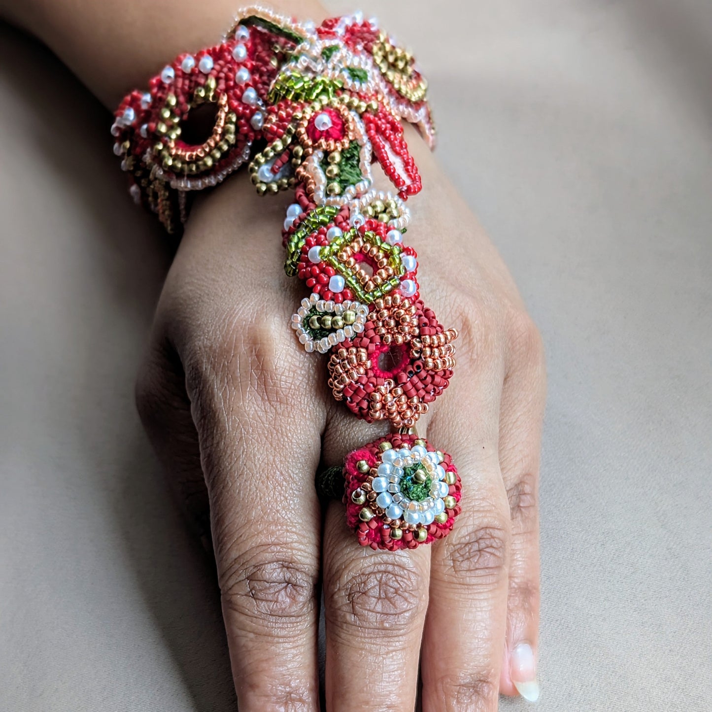 Close up view of a hand modelling the intricately hand crafted Shreya 5-in-1 Indian hand harness bracelet in red with green, gold and pearl accents.