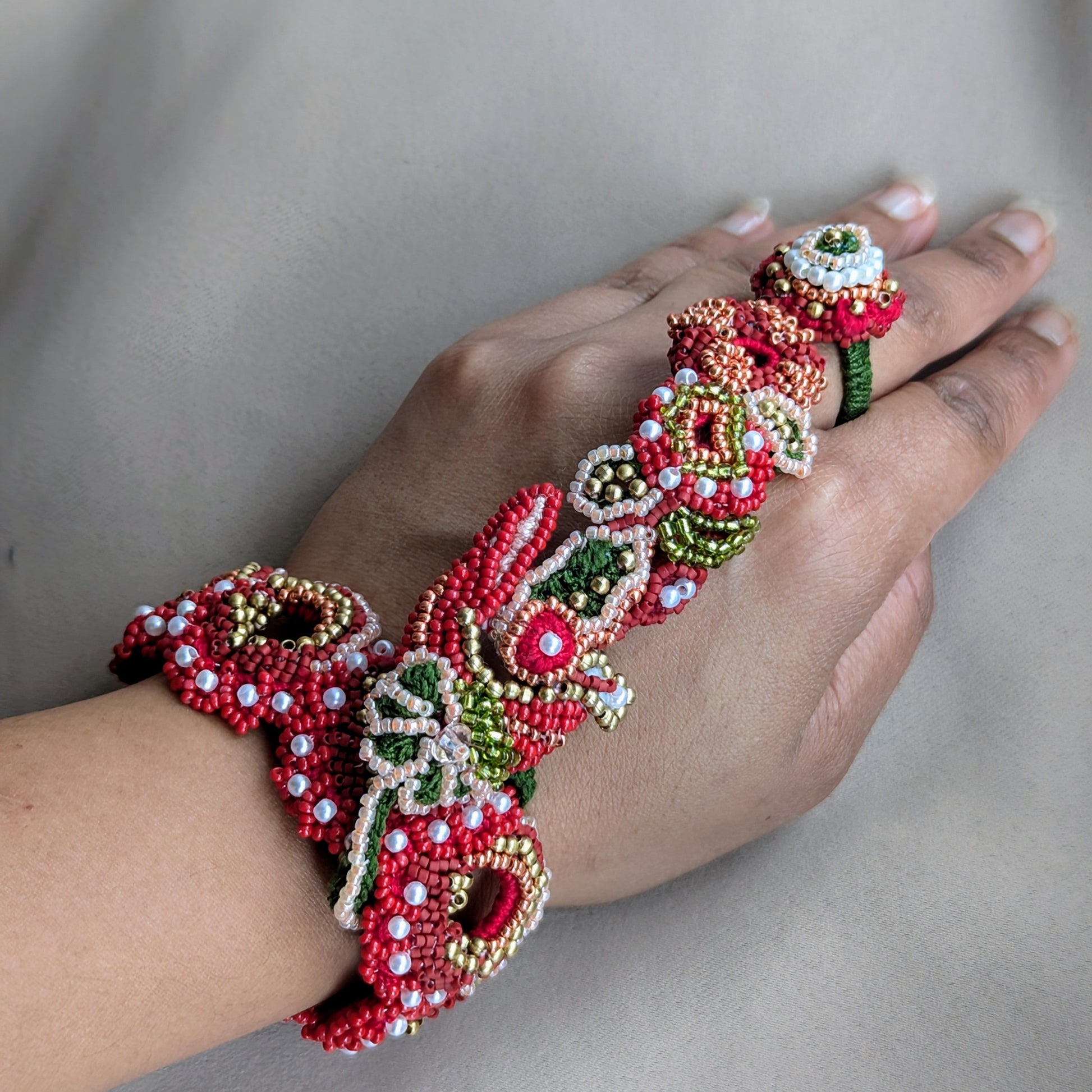 View from above of a hand wearing the hand woven Shreya 5-in-1 Indian haath phool bracelet featuring botanical elements in red and green with gold and pearl accents.