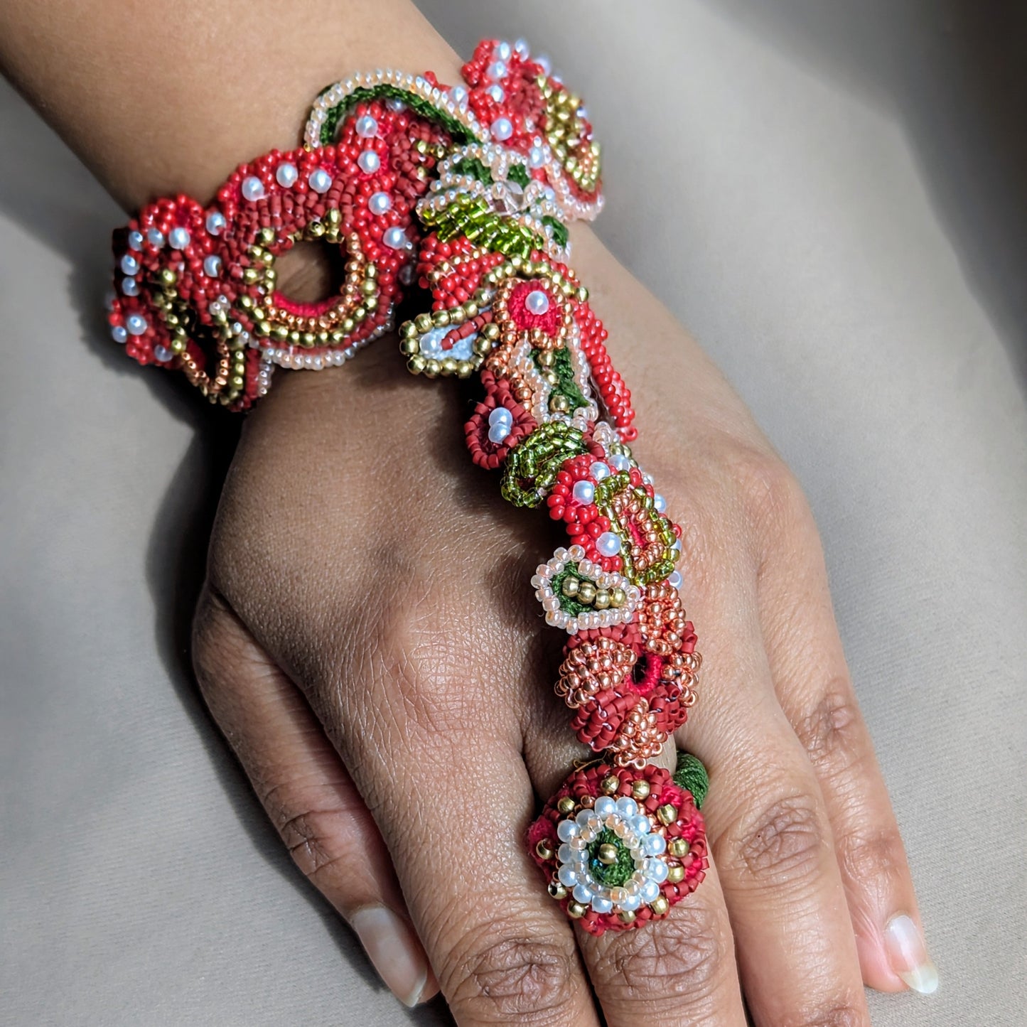 A close-up of a hand wearing the Shreya 5-in-1 highly detailed, ornate beaded jewellery piece that spans from a red beaded wristband down to a large floral ring on the middle finger. The jewellery features red, green, white, and gold seed beads in intricate patterns.