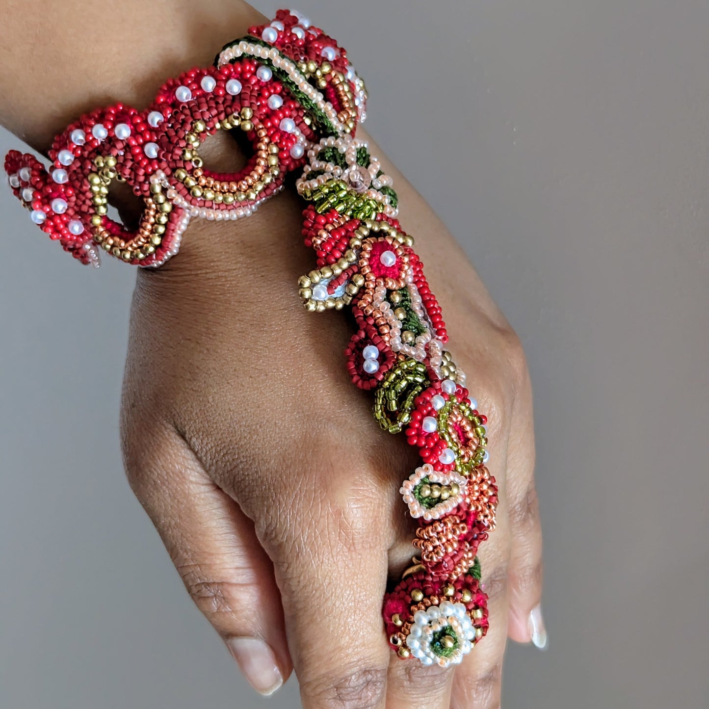 A close up angled view of the Shreya 5-in-1 Indian artisan made luxury haath phool bracelet - featuring red and green with gold and pearl accents - against a neutral grey backdrop.