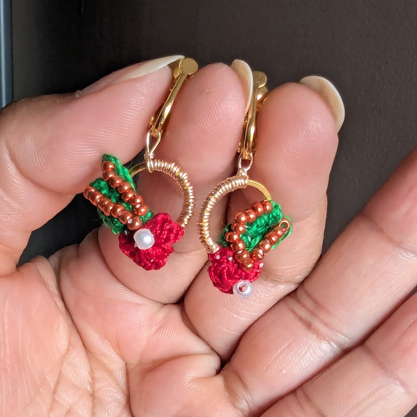 Hand holding an artisan handcrafted Kumkum bead wire crochet pair of comfortable red green gold and copper clip on earrings inspired by nature and Indian heritage style.