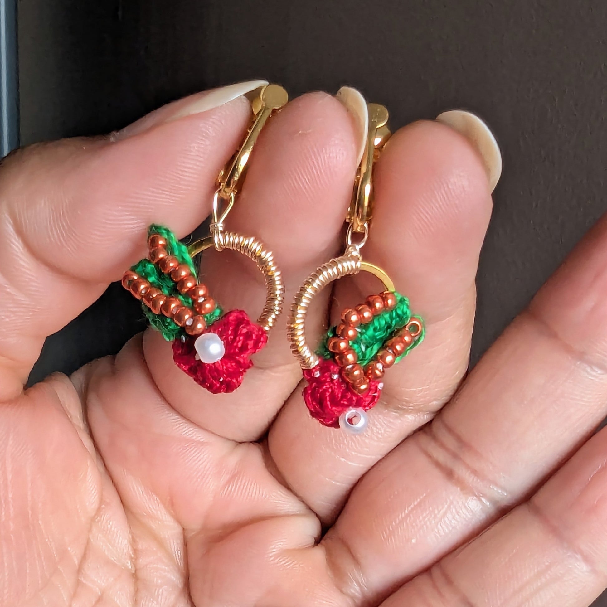Hand holding an artisan handcrafted Kumkum bead wire crochet pair of comfortable red green gold and copper clip on earrings inspired by nature and Indian heritage style.