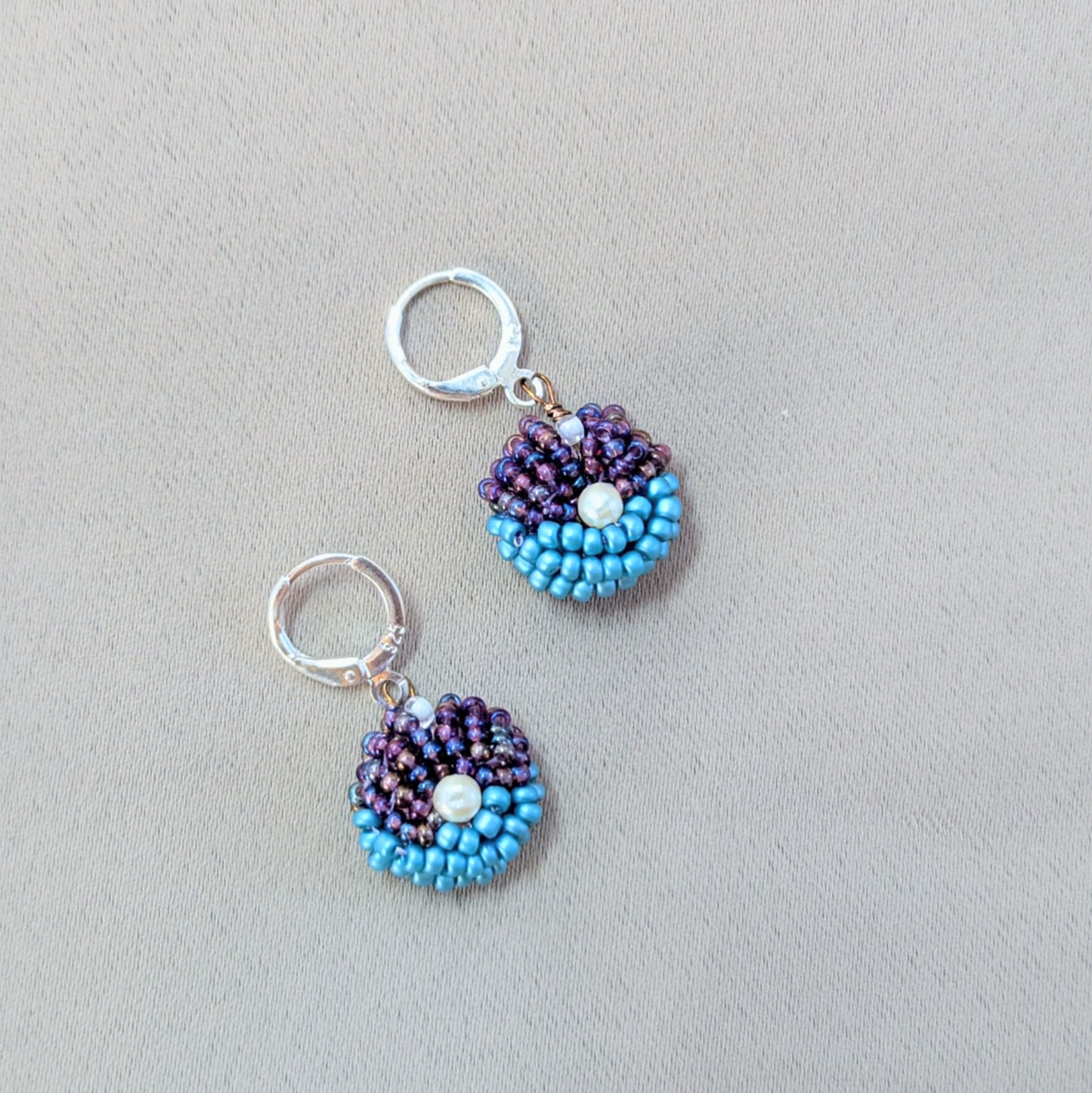 Pair of purple and blue beaded earrings displayed on neutral background showing handcrafted texture and shine.