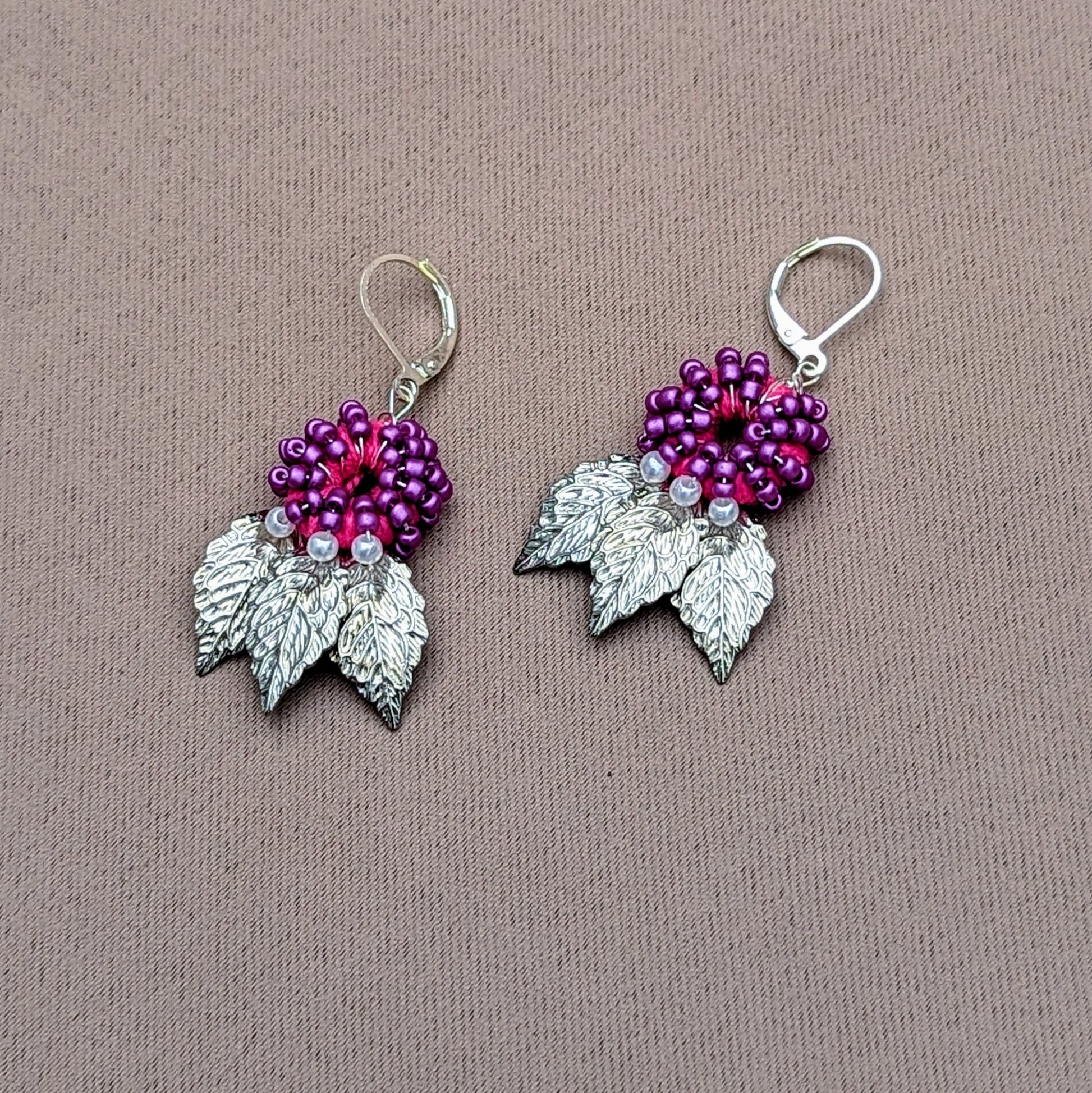 Purple and silver leaf earrings handcrafted with crochet and seed bead detailing on neutral fabric backdrop.