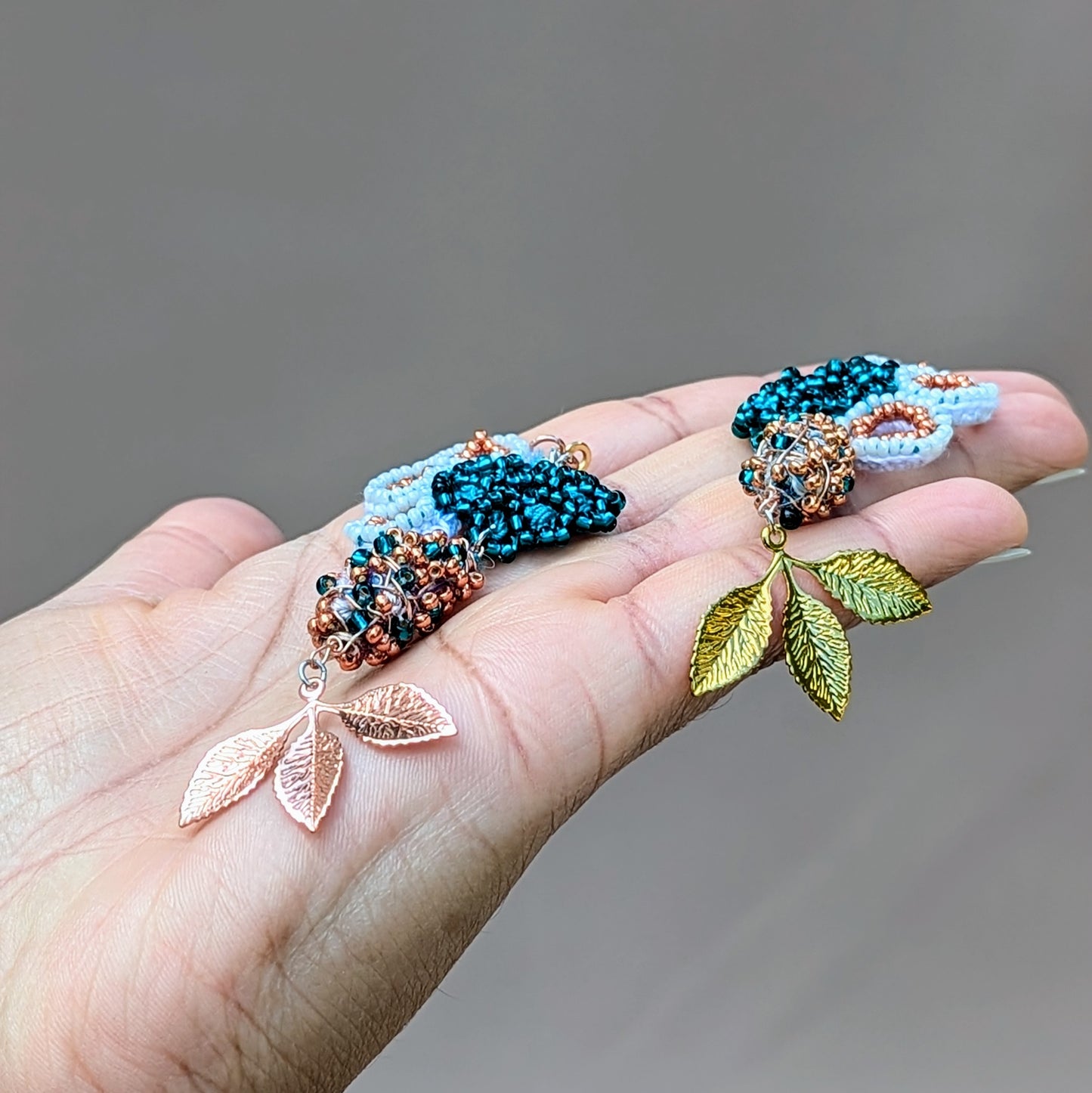 Side angle view of an artisan pair of handmade asymmetric Varuna earrings held on the open palm of a hand. Unique and exclusive to Mayaani Jewellery UK they feature seahorse motifs in bead, crochet and wire work with scrap beads forming the central component ensuring every piece of scrap from the jewellery making process is responsibly repurposed.