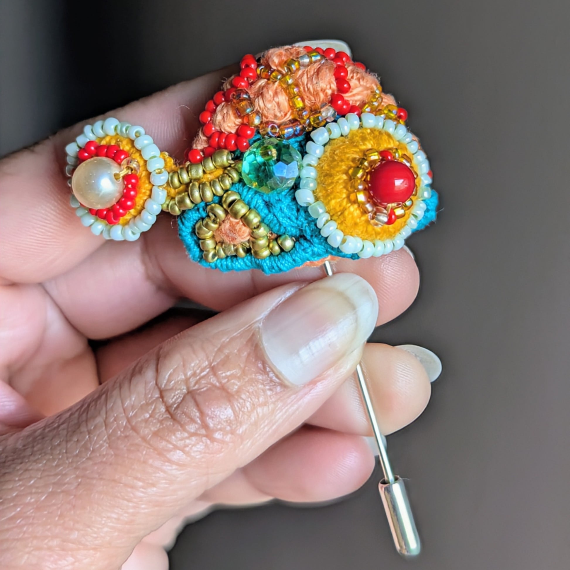 Tejasi handmade beaded brooch in orange, turquoise and red held in hand, featuring vintage crystal glass bead, seed pearls and layered textile embroidery.