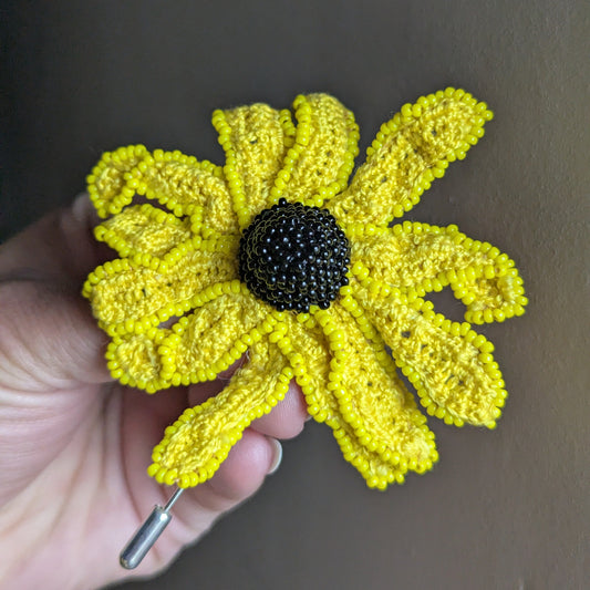 Vamika handmade rudbeckia flower brooch by Mayaani Jewellery -sunshine yellow crochet petals with black seed bead centre held against a grey backdrop.