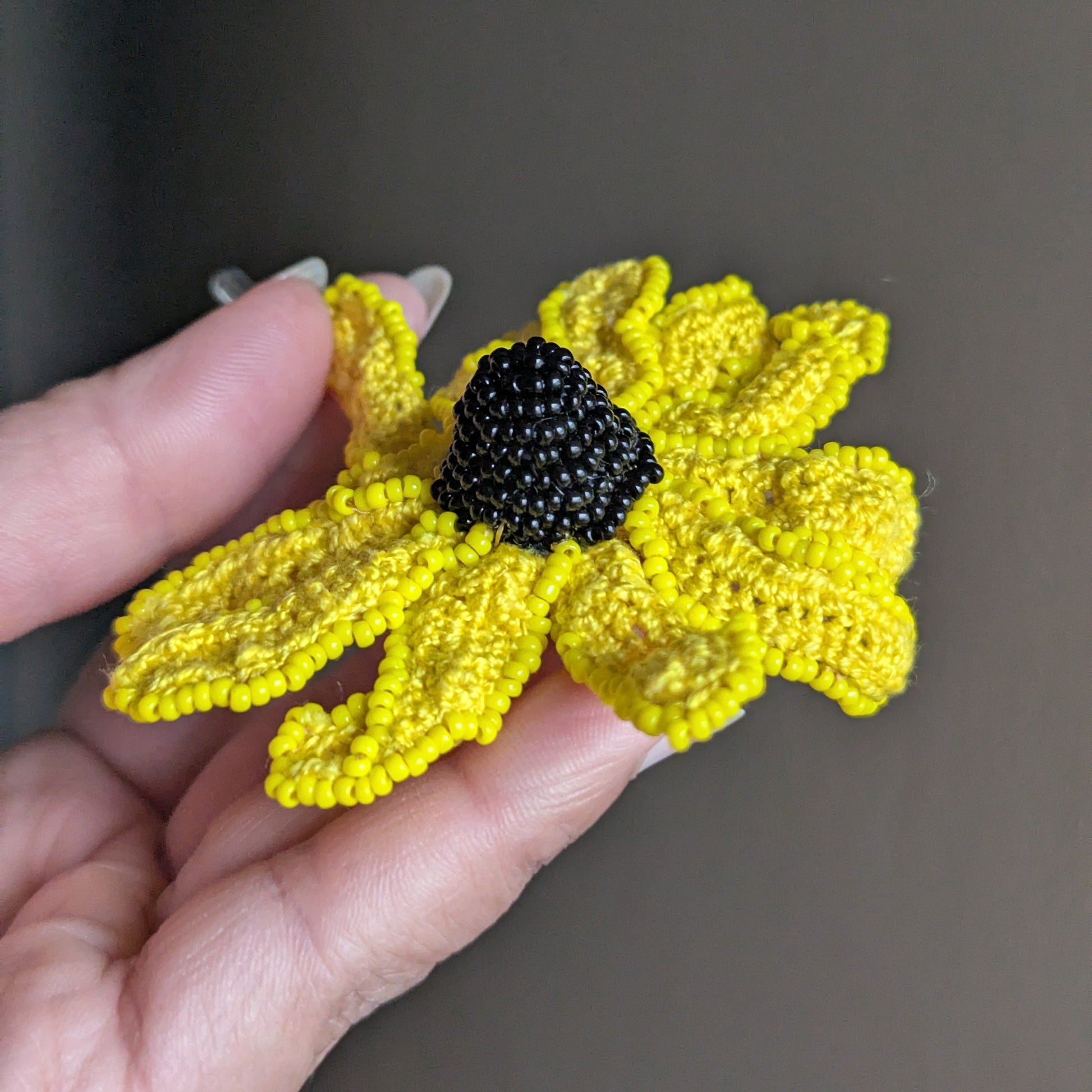 A hand holding the Vamika rudbeckia flower brooch featuring a bold black seed bead centre surrounded by bright yellow cotton petals.