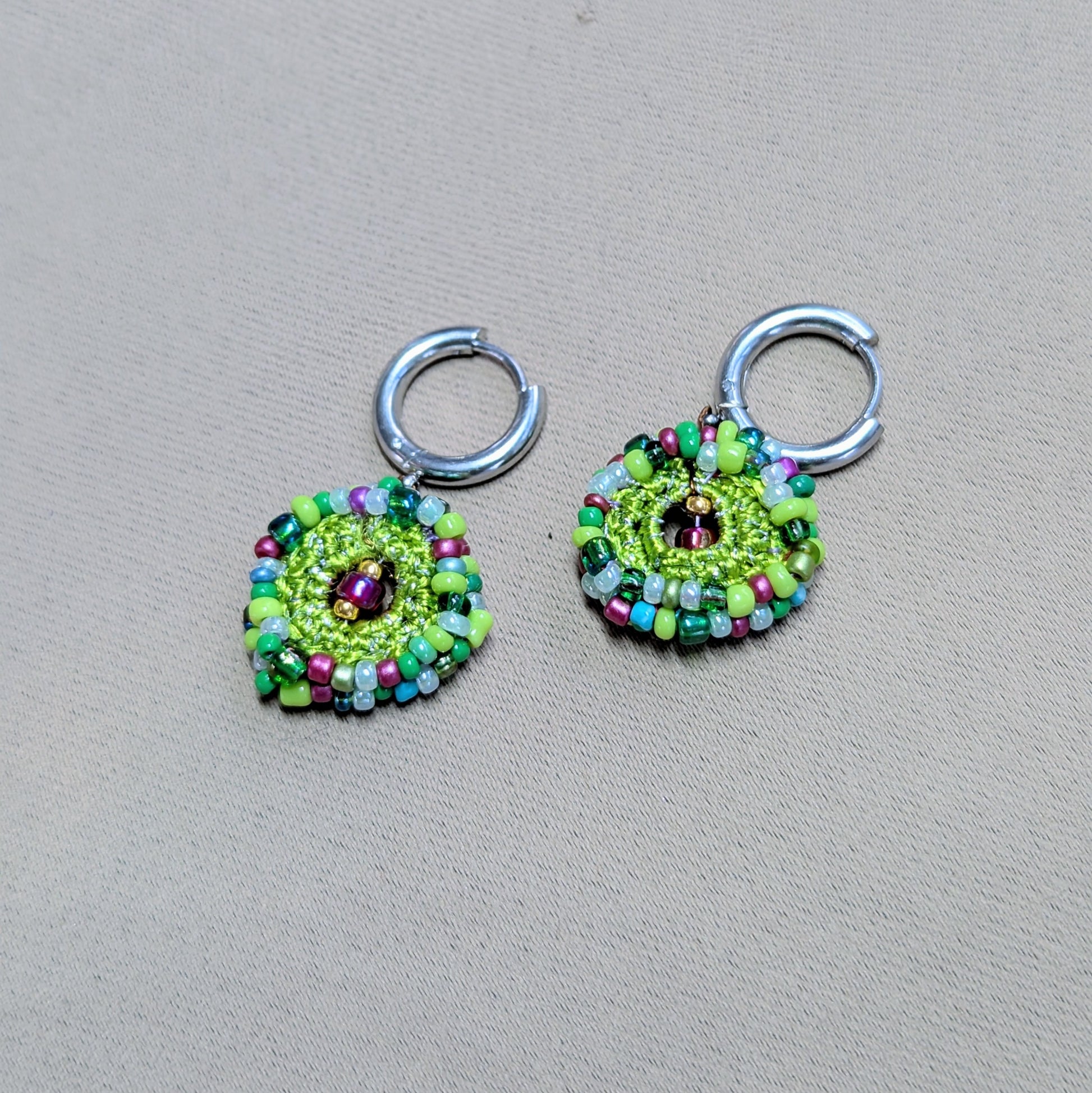 Handcrafted Vana garden-inspired beaded earrings laid flat on a neutral background, featuring green, red, blue, and purple glass seed beads with circular crochet detailing and stainless steel hoop clasps.