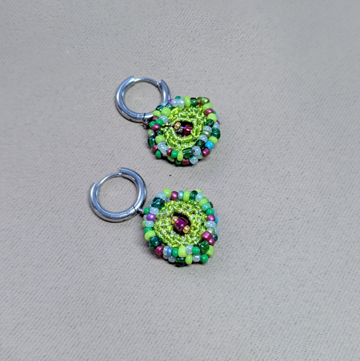 Side-angled lay of Vana artisan bead crochet earrings, showing layered circular construction, mixed glass seed beads, and silver-tone stainless steel hoop findings.