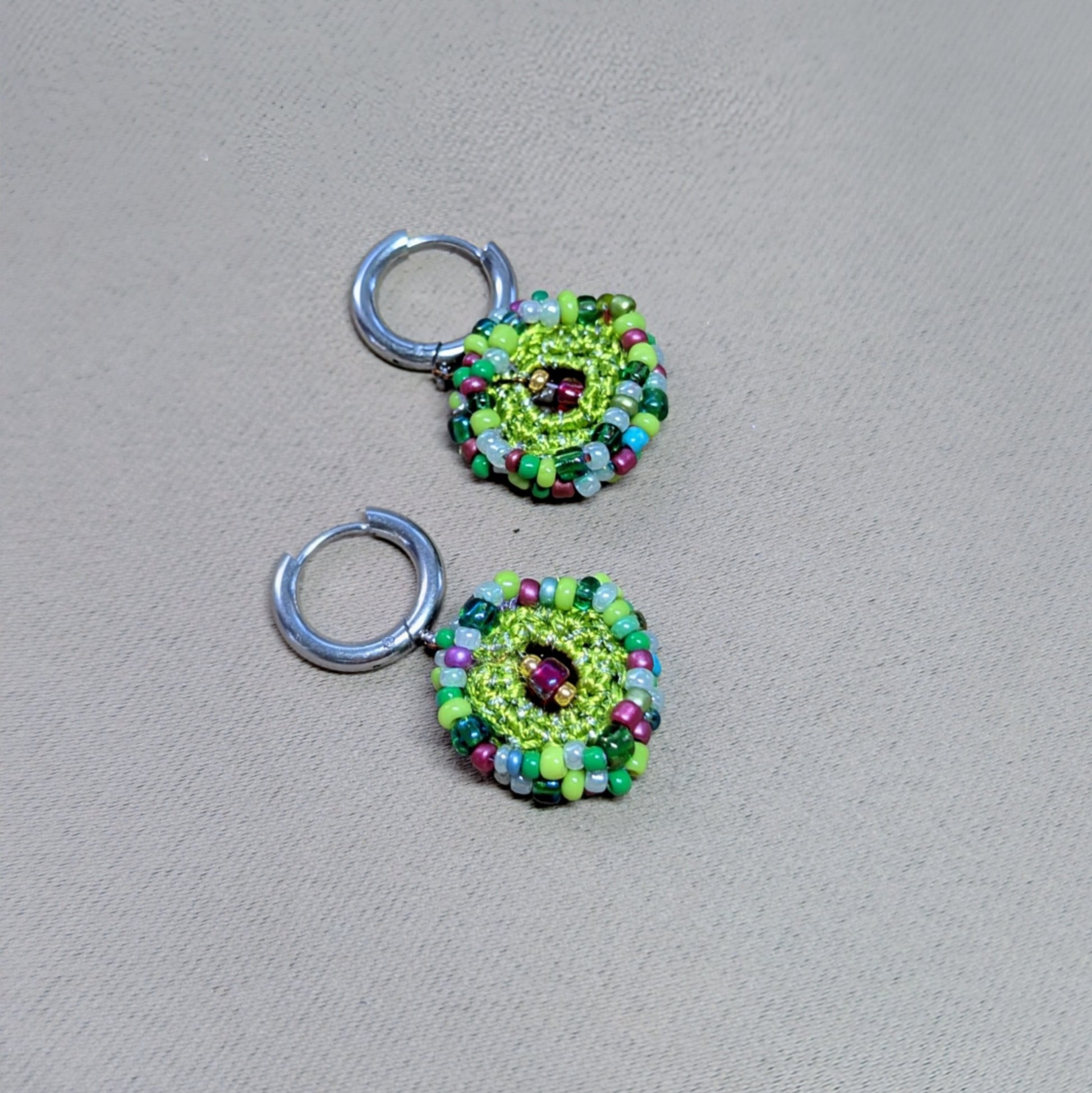 Side-angled lay of Vana artisan bead crochet earrings, showing layered circular construction, mixed glass seed beads, and silver-tone stainless steel hoop findings.