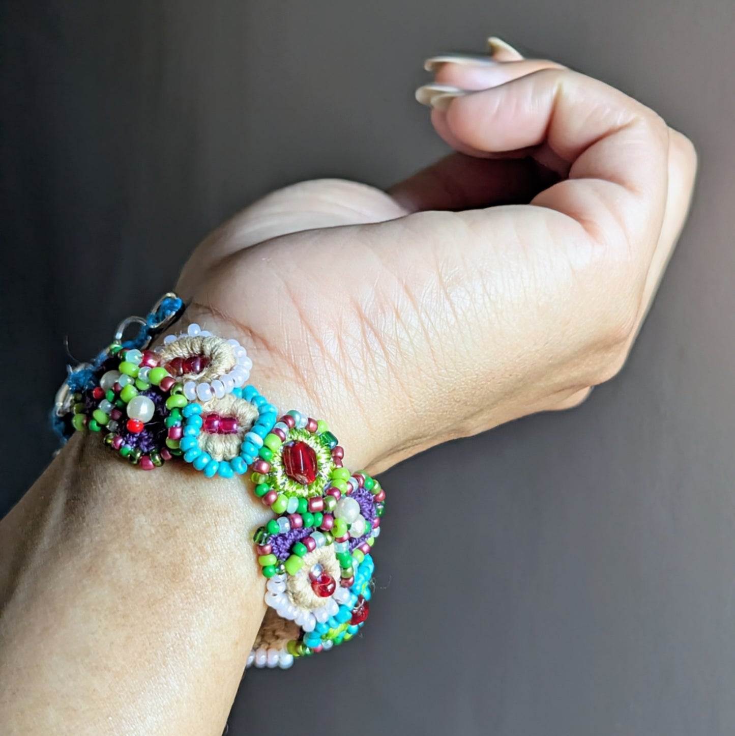 Garden-inspired handmade beaded bracelet shown on the wrist, combining Czech glass seed beads, seed pearls, and cotton thread in green, blue, ivory, and red tones.