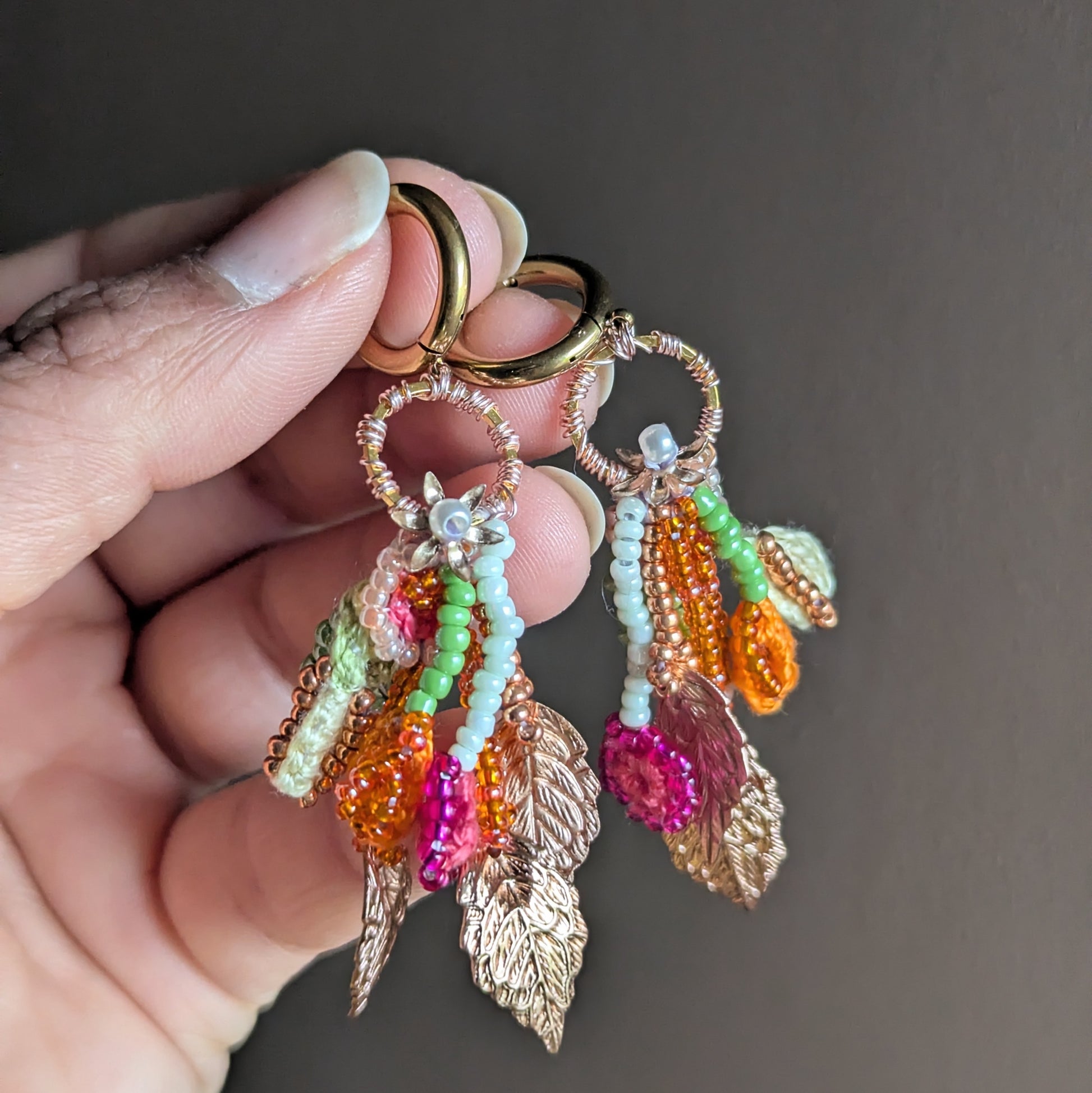 Close-up handheld view of Zariya earrings showing layered bead strands, seed pearls, textile cotton elements and rose gold leaf charms on gold stainless steel hoops.