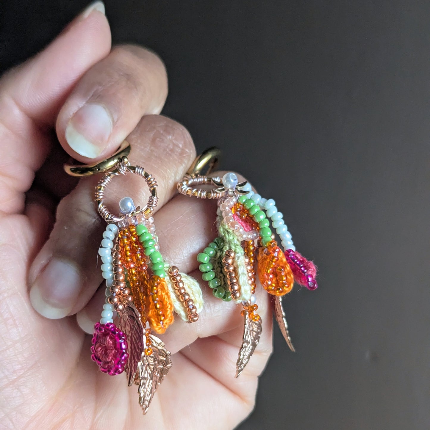 Handheld close-up of Zariya statement earrings with rose gold leaf charms, amber bead drops, green and white seed beads and seed pearl accents, handmade in the UK.