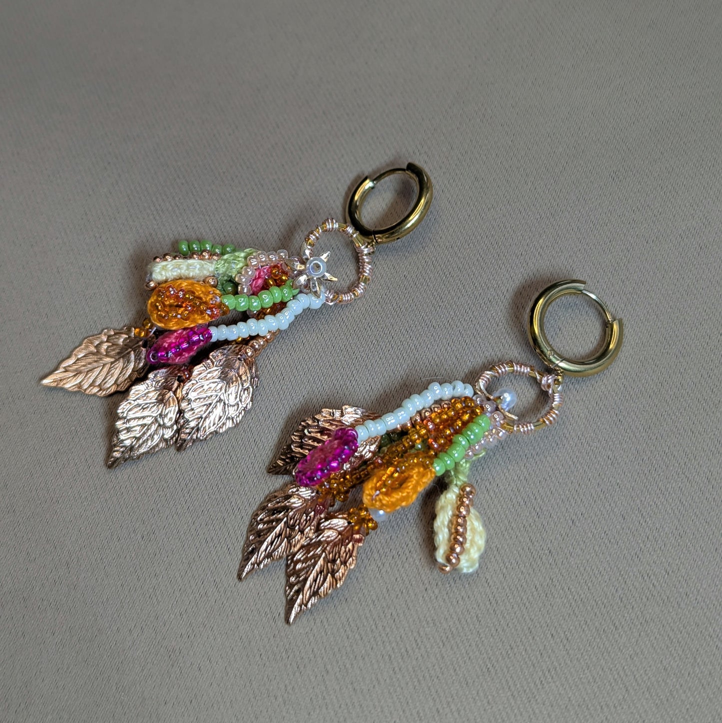 Angled flat lay of Zariya beaded tassel earrings showing rose gold leaf charms, flower detail, seed pearls and mixed Czech glass beads on gold hoops.