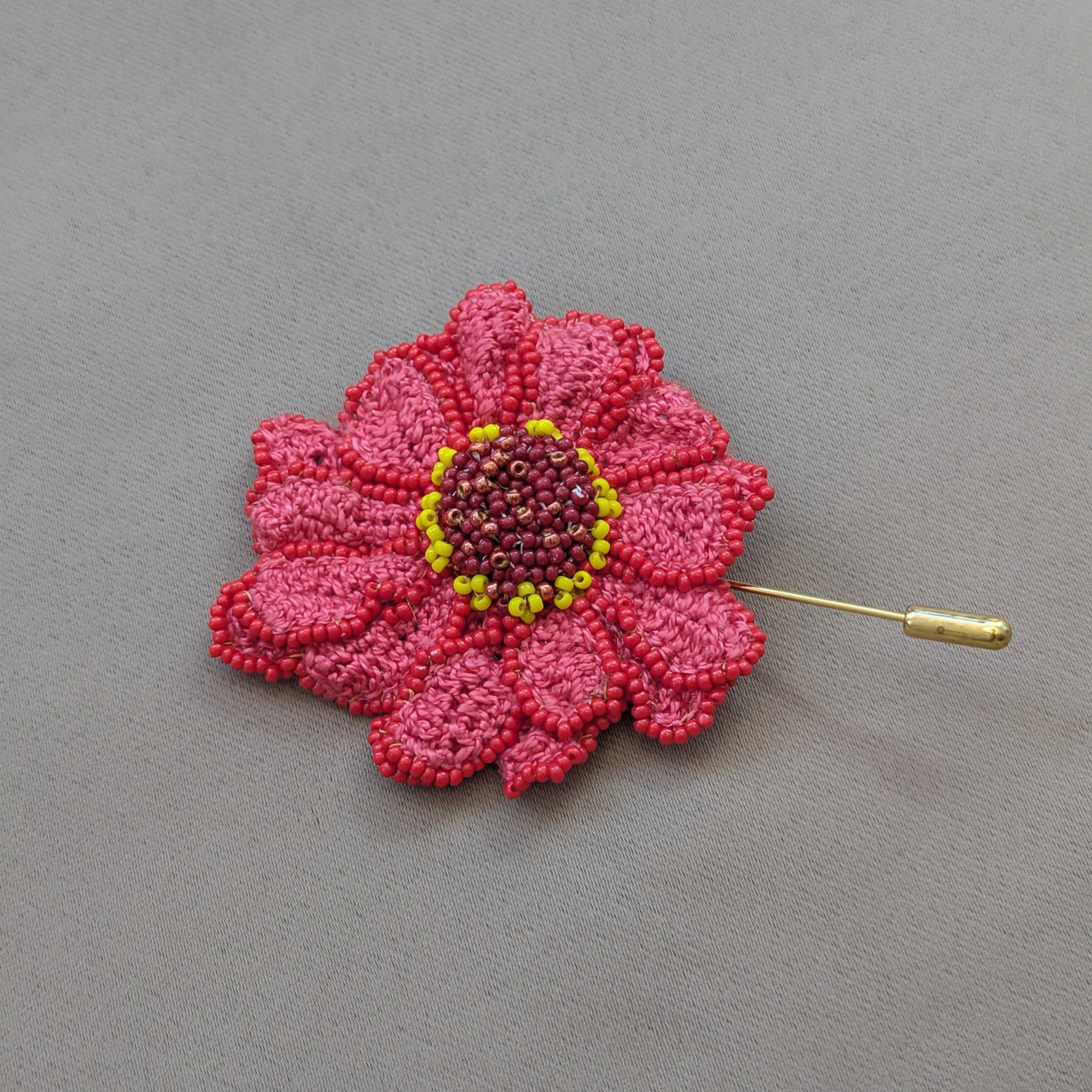 Flat lay view of a textural botanical Zinnia flower brooch in pinks, red and yellow featuring a gold stick pin for wearable art inspired by nature.