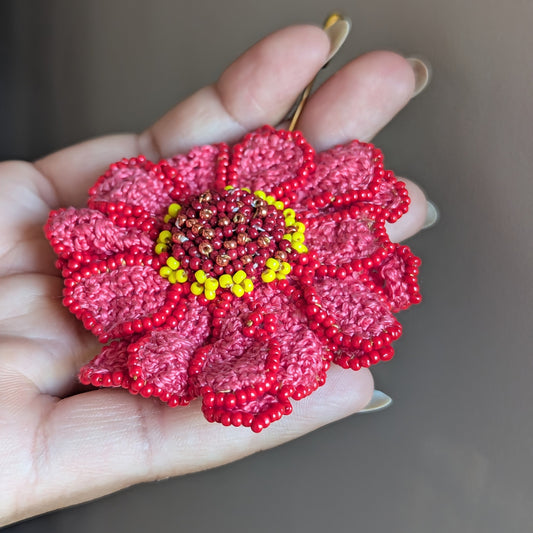 Side view of a large statement zinnia flower lapel, hat, brooch pin featuring red and yellow seed beads edging and embellishing a pink crochet bed of petals. Handcrafted with intention at Mayaani Jewellery UK.
