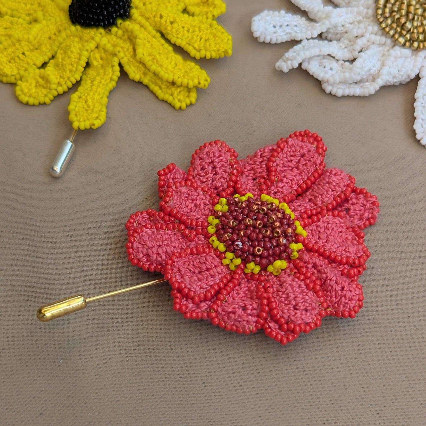 A one off pink Zinnia flower pin pictured on a neutral grey backdrop with a white Daisy and yellow Rudbeckia brooch pin visible in the backdrop - part of the Garden of Becoming collection from Mayaani Jewellery UK.